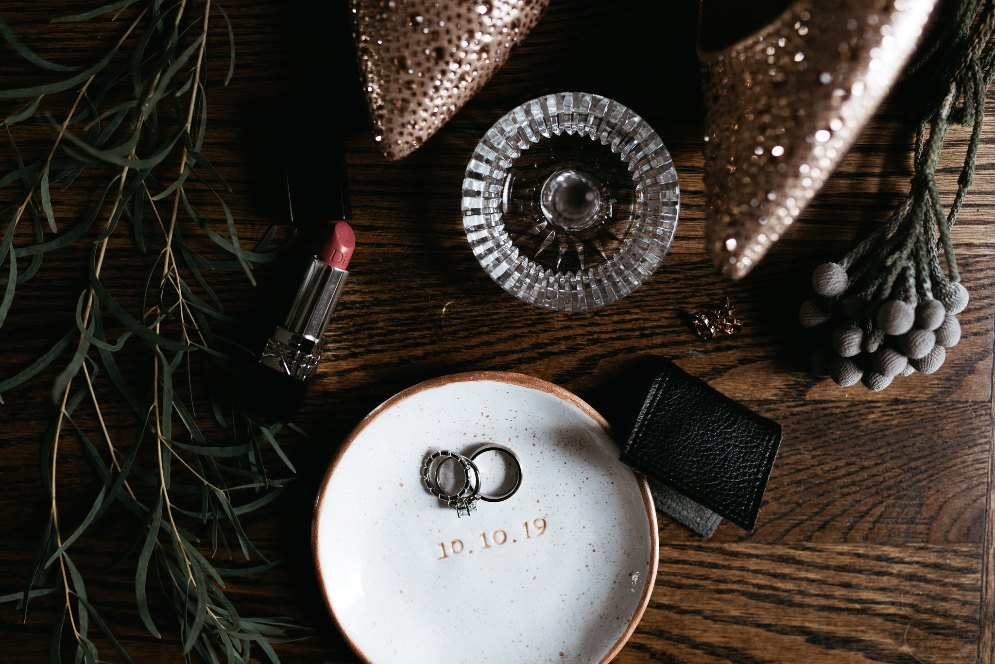 Moody photograph of romantic wedding details featuring gold rhinestone heels, Dior lipstick, a handmade ring dish with wedding rings on it, greenery from the bouquet and small gold earrings.
